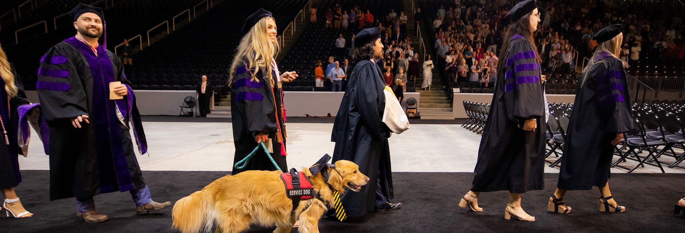 Students walk into Thompson-Boling Arena at commencement, one student with a golden retriever by her side wearing a red vest labeled "service dog."